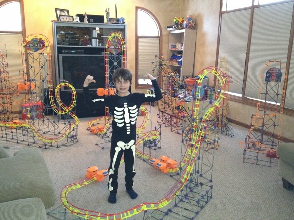 Young Aiden in a skeleton costume, surrounded by a living room full of K’NEX roller coasters with loops, towers and winding track.